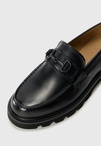 Black leather loafer with a smooth finish, a decorative strap and metallic accent, rounded toe, and textured rubber sole.