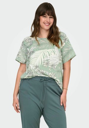 Femme souriante portant une chemise à manches courtes à motif tropical de feuilles vertes et de fleurs, rentrée dans un pantalon sarouel vert sarcelle à cordon de serrage.
