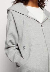 Kvinna som bär en ljusgrå zip-up hoodie med framficka och vit innerskjorta, handen i fickan, inget synligt ansikte.
