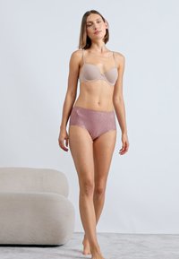 Kvinde står barfodet iført en beige BH og mauve højtaljede blondeundertøj mod en simpel lys baggrund.