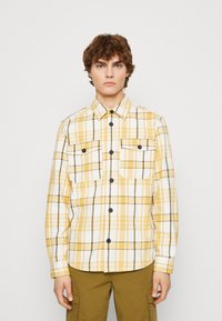 Chemise à carreaux jaune et blanc avec des accents noirs, deux poches avant, fermeture à boutons et manches longues. Fabriquée à partir d'un tissu texturé.