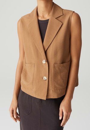 Sleeveless brown blazer with two buttons and front pockets worn over a black top, paired with a dark skirt.