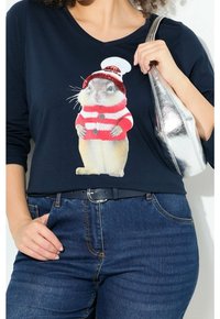 Frau in einem marineblauen V-Ausschnitt-Shirt, auf dem ein Eichhörnchen abgebildet ist, das einen rot-weißen gestreiften Pullover und eine Mütze trägt, kombiniert mit blauen Jeans und einer silbernen Tasche.