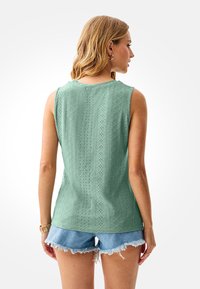 Green sleeveless top with textured diamond pattern; round neckline; paired with light blue frayed denim shorts, worn from the back.