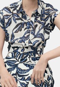 Femme portant une chemise à manches courtes boutonnée et une jupe assortie avec des motifs de feuilles tropicales noires et bleues sur fond blanc.