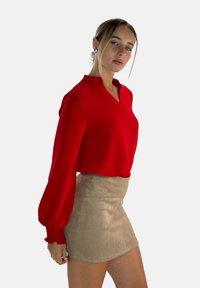 Red, lightweight, long-sleeve top with a collar and gathered cuffs, paired with a fitted beige suede mini skirt. Smooth texture, solid colors.