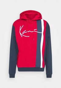 Sudadera con capucha de frente rojo, mangas azul marino, rayas verticales azul marino, blancas y rojas a la derecha, y logo en estilo firma en blanco en el pecho.