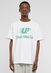 Balta medvilninė marškinėliai trumpomis rankovėmis su laisvu pjūviu. Pabrėžia žalią grafiškai atvaizduotą tekstą "Upscale Athletic Club" ir logotipą.