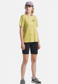 Maglietta a maniche corte di colore giallo limone con una grafica sul petto, abbinata a pantaloncini neri. Il modello indossa un berretto grigio chiaro e scarpe da trekking grigie.