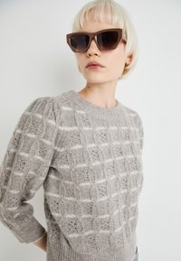 Pull en tricot gris clair avec un motif texturé de côtes et de ajourés, présentant un col rond et de longues manches. Lunettes de soleil oversized marron.
