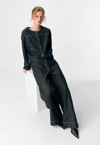 Schwarzes gestreiftes Stoffoutfit mit einem geknöpften Oberteil und weitem Hosenbein. Mit passendem Nähen und spitzen schwarzen Schuhen.
