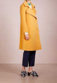 Cappotto di lana giallo con ampio colletto, bottoni doppi, e una silhouette rilassata, abbinato a pantaloni cropped scuri e mules floreali.
