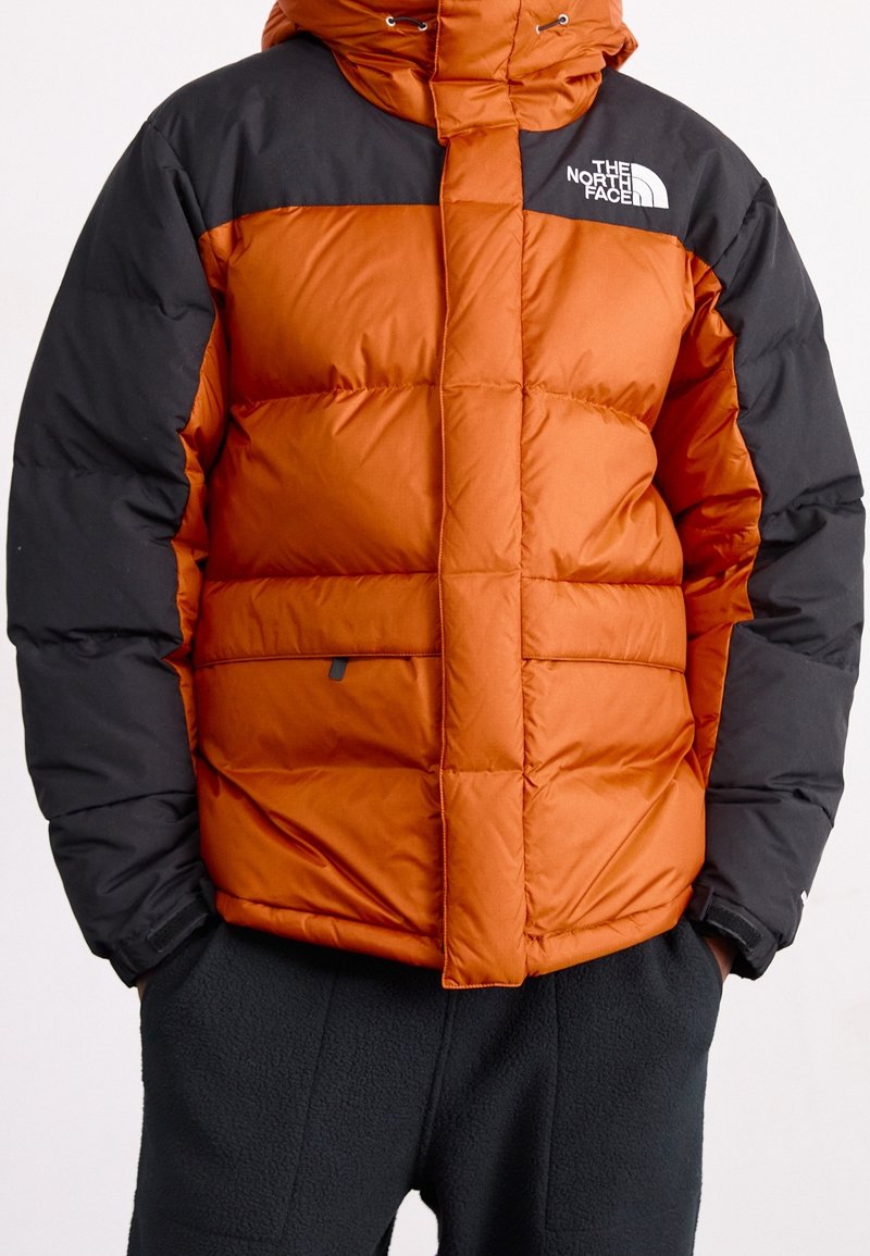 Doudoune orange et noire avec capuche, dotée de sections matelassées, d'une fermeture éclair à l'avant et d'accents de logo sur la poitrine et la manche.