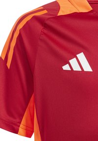 Rode sportshirt met oranje accenten, korte mouwen en een witte logo. De stof lijkt glad met een lichte textuur en een sportief ontwerp.