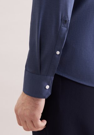 Mands hånd ved siden af navyblå langærmet skjortemanchet med hvide knapper og broderet sort rosedetalje på manchetstrop.