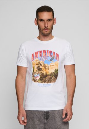 Homme portant un T-shirt blanc avec un motif coloré de Mount Rushmore et d'aigle avec le texte « American Life », se tenant devant un fond uni.