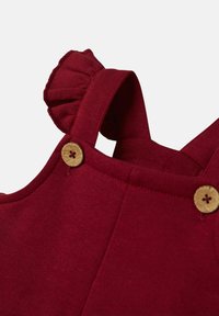 Rote Baumwoll-Latzhose mit strukturierten Trägern und Holzknöpfen. Verfügt über einen ausladenden Rüschensaum an der Schulter und ein glattes, enganliegendes Design.