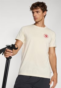 Krémové tričko s krátkým rukávem a kulatým výstřihem, s červenou nášivkou loga na levé hrudi. Model drží černou turistickou hůl.