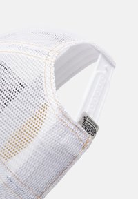 Bílá síťovaná kšiltovka se zlatým prošíváním, nastavitelným plastovým popruhem a značkovou etiketou. Disponuje prodyšným designem a geometrickým vzorem.