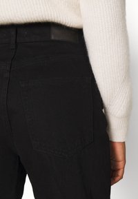 Pantalones de mezclilla negros con dos bolsillos traseros, con una cintura texturizada y etiqueta de cuero; visión cercana de la parte trasera.