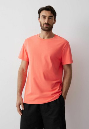 Mężczyzna z ciemnymi włosami i brodą, ubrany w jasnoróżową koszulkę z krótkim rękawem i czarne szorty, stojący na tle jednolitego jasnego tła.