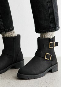 Czarne botki za kostkę z dwoma złotymi klamrami, noszone z jasnymi skarpetami dzianinowymi i czarnymi dżinsami na szarym podłożu i białej ścianie.