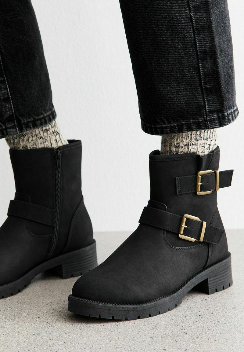 Botas negras hasta el tobillo con dos hebillas doradas, llevadas con calcetines de punto claros y pantalones de denim negros, sobre un suelo gris y una pared blanca.