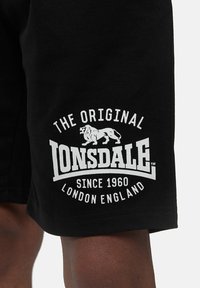 Melni kokvilnas īsi šorti ar baltu izdrukātu logotipu, uz kura ir lauva un uzraksts "ORIGINAL LONSDALE SINCE 1960 LONDON ENGLAND."
