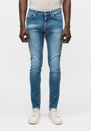 Obcisłe niebieskie dżinsy noszone z białymi trampkami na stojącej osobie, pokazane od pasa do stóp na jednolitym tle.