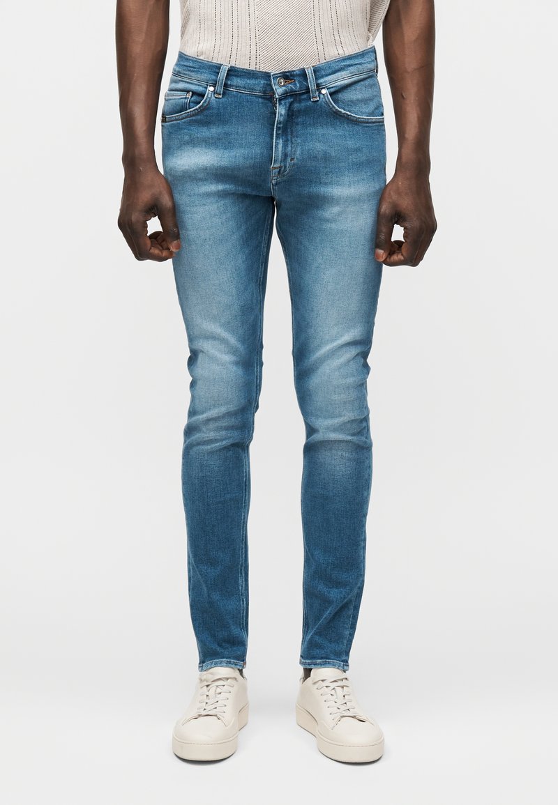 Στενά μπλε τζιν φοράει ένα άτομο που στέκεται, με λευκά αθλητικά παπούτσια, εμφανίζεται από τη μέση έως τα πόδια σε απλό φόντο.