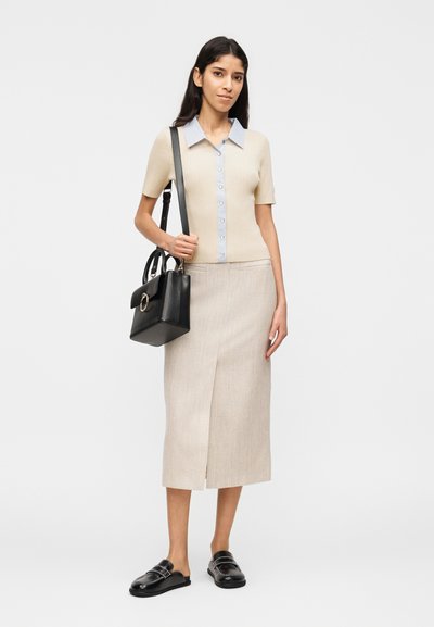 Γυναίκα που φοράει μπεζ κοντομάνικο πουκάμισο με κουμπιά, μπεζ μίντι φούστα με άνοιγμα εμπρός, μαύρα μοκασίνια και κρατάει μαύρη τσάντα ώμου.