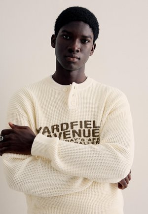 Mladý muž s krátkymi kučeravými vlasmi, oblečený v krémovom svetri so vzorom vafle a nápisom „Yardfield Avenue“, so založenými rukami, neutrálne pozadie.