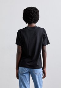 Osoba s krátkými, kudrnatými vlasy nosí černé tričko a modré džíny, stojí před jednobarevným pozadím.