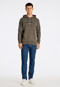 Felpa verde oliva con lacci e tasca frontale, abbinata a jeans blu e sneakers bianche. Design semplice, texture morbida.