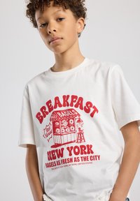 White cotton t-shirt featuring a bold red graphic of a bagel stand with text "BREAKFAST NEW YORK" and "BAGELS AS FRESH AS THE CITY."