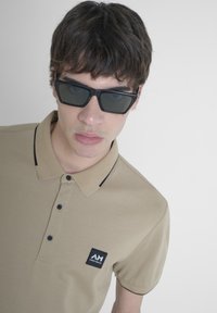 Beige polo shirt met een geribde kraag, twee knopen, zwarte accenten en een vierkante logopatch op de borst. Draag het met donkere zonnebrillen.