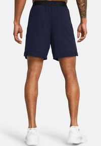 Mann in marineblauen Sportshorts und weißen Turnschuhen, von hinten betrachtet, mit entspannten Armen an den Seiten.