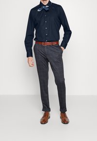 Donkerblauwe overhemdd met knoopjes, grijs geruite stijlvolle broek, bruine leren riem en bruine nette schoenen. Het shirt heeft een halsdoekdetail.