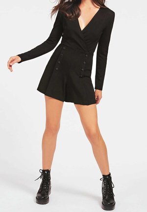 Schwarzer Langarm-Overall mit V-Ausschnitt, Knopfdetails auf der Vorderseite und A-Linien-Shorts. Kombiniert mit schwarzen Schnürstiefeln im Combat-Stil.