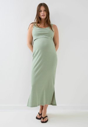 Femeie însărcinată în picioare, purtând o rochie lungă fără mâneci cu dungi verzi și albe și șlapi negri, pe un fundal alb simplu.