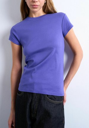 Woman wearing a fitted short-sleeve purple t-shirt and black jeans, standing against a plain light-colored wall.