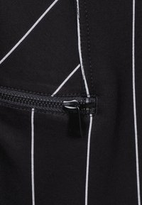 Tissu noir avec des rayures blanches verticales, comportant une petite poche zippée mise en valeur par une fermeture éclair noire contrastante.