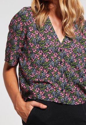 Vrouw draagt een korte mouw V-hals blouse met een roze, groene en witte bloemenprint en zwarte broek met één hand in de zak.