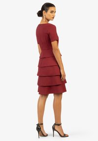 Bordeaux-farbenes, gestuftes Kleid mit kurzen Ärmeln, Peplum-Taille, Reißverschluss auf der Rückseite und geschichteten Rüschen. Getragen mit schwarzen Riemchen-Pumps.