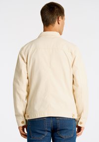 Beige bomullskavaj med klassisk krage, knappade ärmslut och ryggsöm. Har en rak nederkant och sidjusteringar med två knappar.