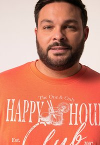 Muž s vousy, který má na sobě oranžové tričko s bílým textem a ilustrací nápoje, na kterém je napsáno "Jedinečný a pouze Klub Šťastné Hodiny Založen 2007."