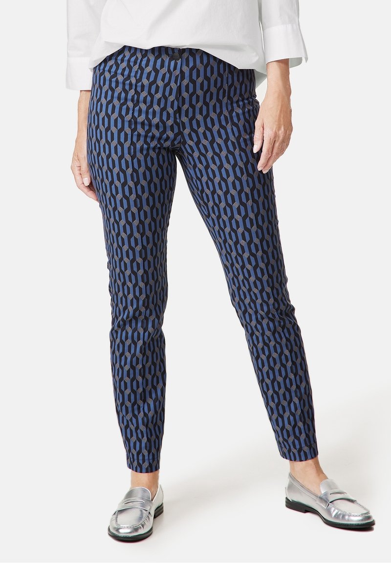 Femme portant un pantalon bleu marine slim avec un motif géométrique noir, un haut blanc et des mocassins argentés, debout sur fond blanc.