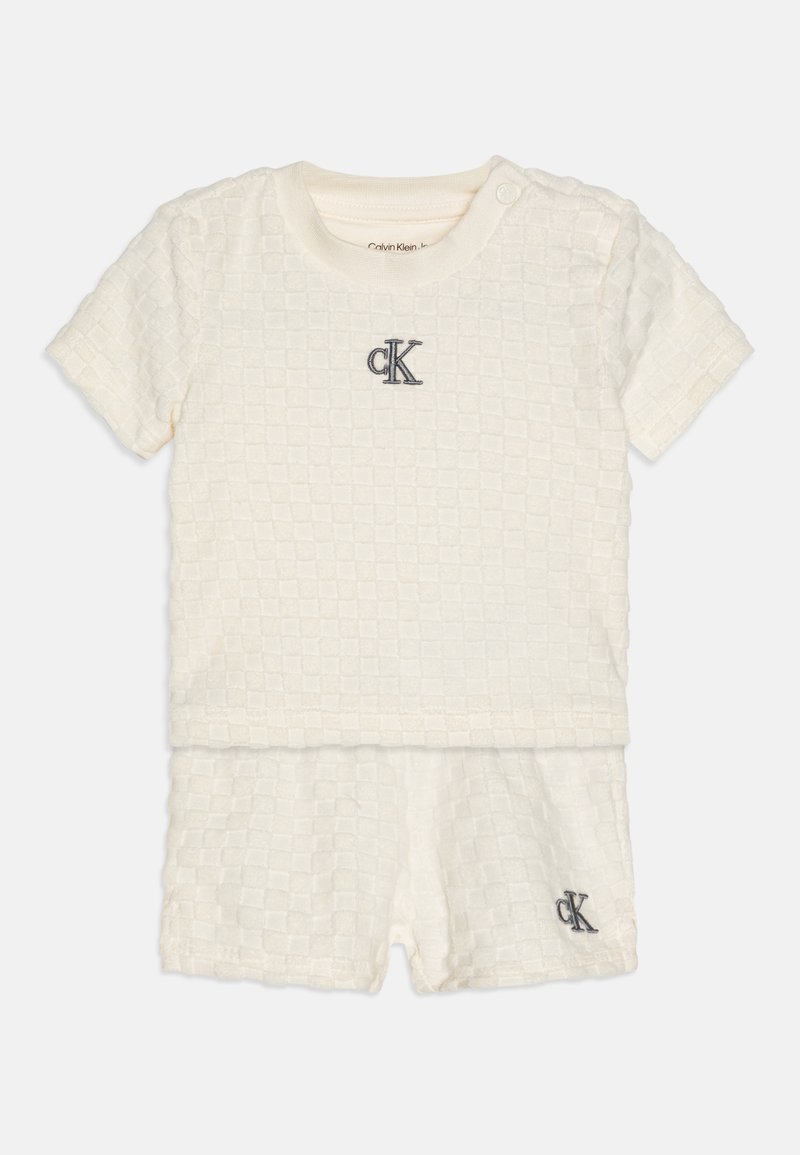 Kortærmet, cremet to-delt sæt lavet af tekstureret stof. Har et præget firkantet mønster og broderet "CK" logo i marineblå.