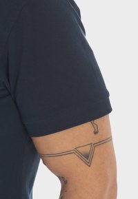 Camicia corta a maniche con tessuto a trama in colore navy. Presenta un collo a giro e una vestibilità aderente, che mette in risalto un tatuaggio sul braccio.