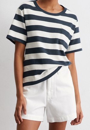 Personne portant un t-shirt à manches courtes rayé horizontalement noir et blanc avec une poche devant et un short blanc taille haute.